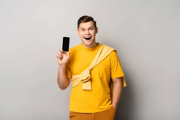 Positive man in yellow t-shirt holding smartphone with blank screen on grey background — Stock Photo