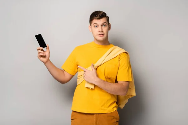 Hombre excitado apuntando con el dedo al teléfono móvil con pantalla en blanco sobre fondo gris - foto de stock