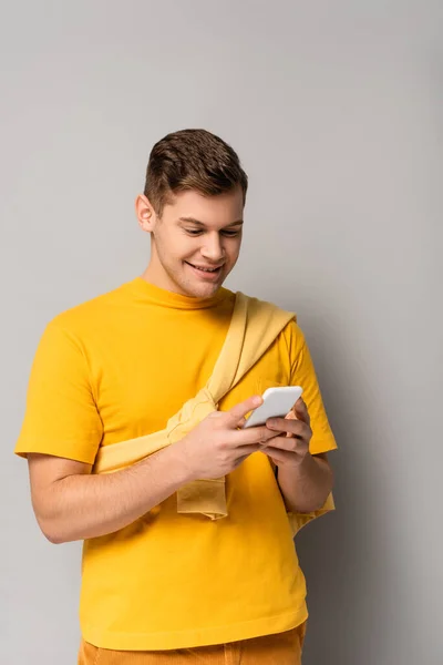 Homem sorridente em camiseta amarela usando smartphone em fundo cinza — Fotografia de Stock