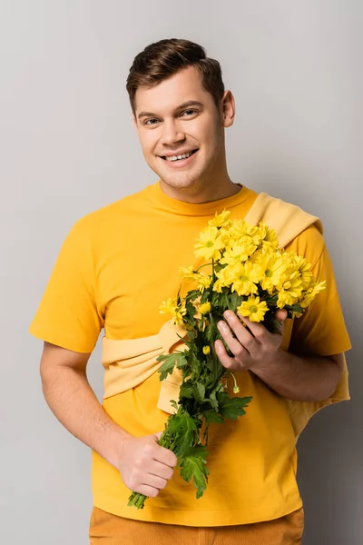 Young man smiling at camera while holding yellow chrysanthemums on grey background — Stock Photo