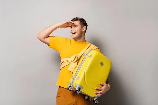 Smiling tourist with yellow suitcase looking away on grey background — Stock Photo