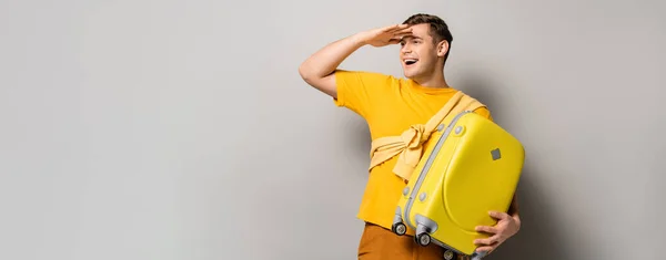 Cheerful tourist looking away while holding yellow suitcase on grey background, banner — Stock Photo