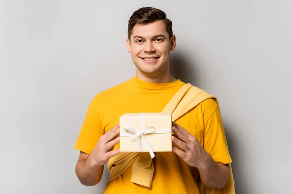 Happy brunette man holding gift with ribbon on grey background — Stock Photo
