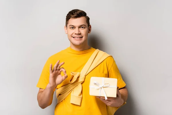Молодой человек с подарочной коробкой показывает нормальный жест на сером фоне — стоковое фото