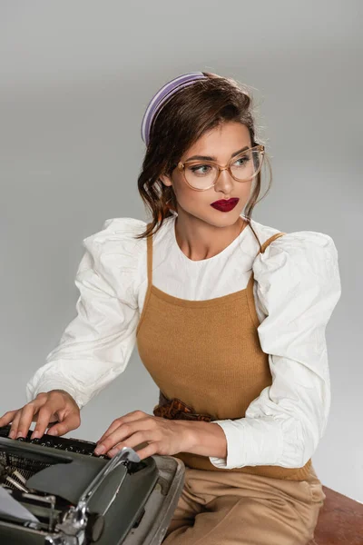 Trendige Sekretärin, die wegschaut, während sie auf einer alten Schreibmaschine tippt, isoliert auf grau — Stockfoto