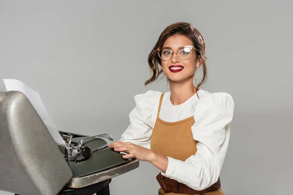 Cheerful secretary typing on retro typewriter and smiling at camera isolated on grey — Stock Photo