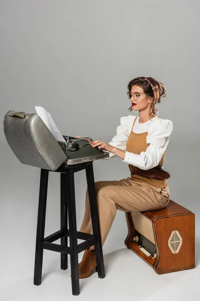 Secretary in retro clothes sitting on radio receiver and typing on vintage typewriter on grey — Stock Photo