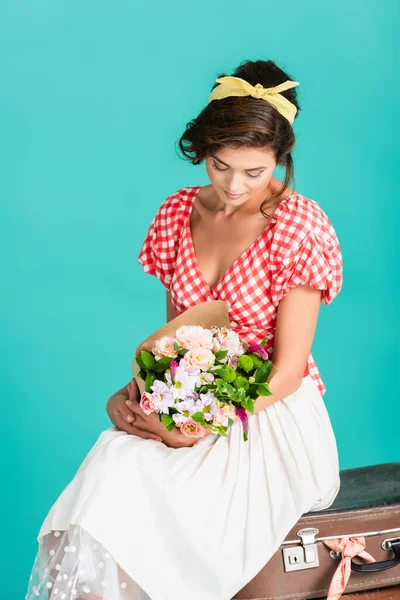 Élégant pin up femme tenant des fleurs tout en étant assis sur une valise vintage isolé sur turquoise — Photo de stock
