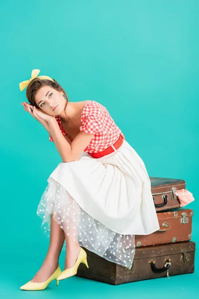 Thoughtful pin up woman holding hands near face while sitting on vintage suitcases on turquoise — Stock Photo