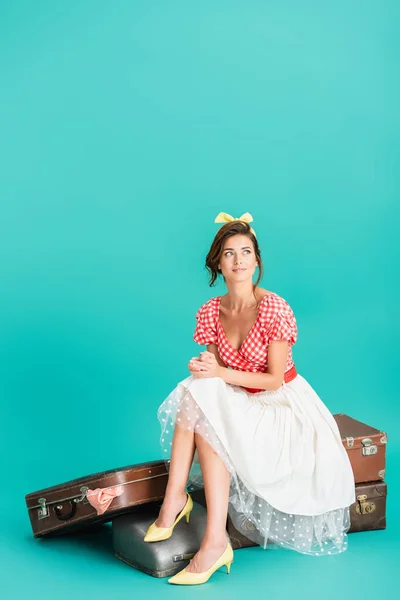 Smiling pin up woman looking away while sitting on vintage suitcases on turquoise — Stock Photo