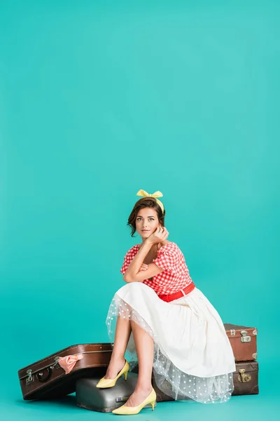 Pretty pin up woman sitting on vintage suitcases and looking at camera on turquoise — Stock Photo