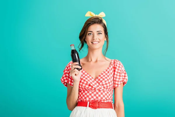 Счастливая женщина в модной ретро-одежде улыбается в камеру, держа бутылку газировки изолированной на бирюзовой — стоковое фото