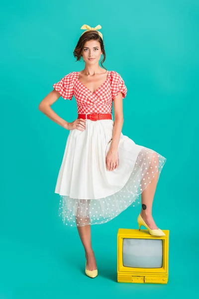 Elegante pin up donna con mano sul fianco guardando la fotocamera mentre calpestando giallo vintage tv su turchese — Foto stock