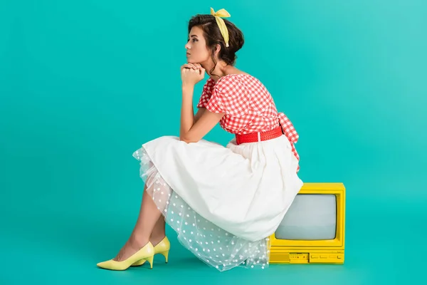 Vue latérale de rêveuse pin up femme assise sur un téléviseur vintage jaune sur turquoise — Photo de stock