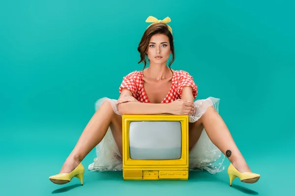 Junge Pin-up-Frau blickt in die Kamera, während sie in der Nähe knallgelber Vintage-Fernseher auf Türkis sitzt — Stockfoto