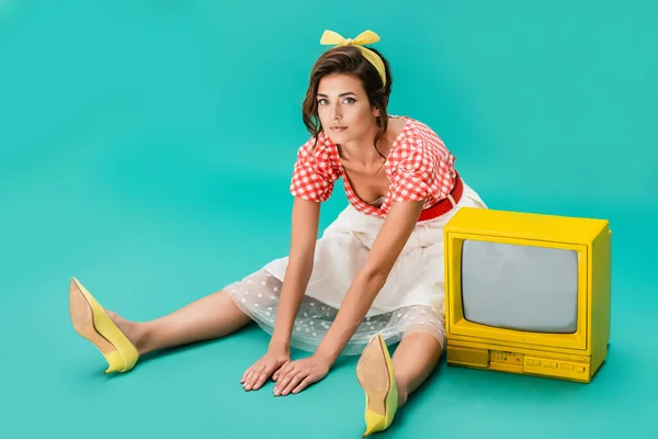 Pretty pin up woman sitting on floor near bright yellow vintage tv and looking at camera on turquoise — Stock Photo