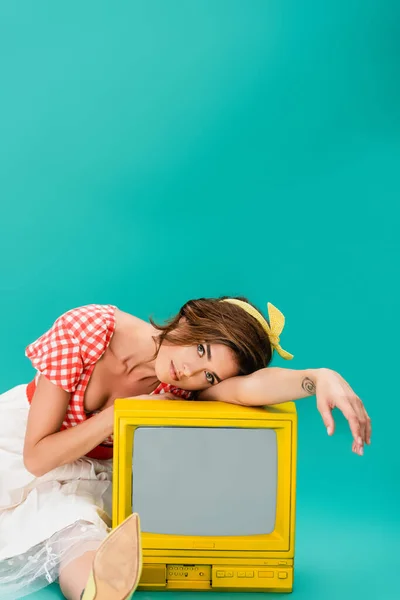 Femme à la mode en vêtements vintage regardant la caméra tout en s'appuyant sur la télévision vintage jaune sur turquoise — Photo de stock