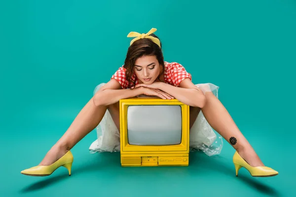 Stylish pin up woman leaning on yellow retro tv set while sitting on turquoise background — Stock Photo