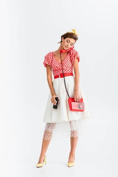 Full length view of offended pin up woman with bottle of soda talking on vintage phone on white — Stock Photo