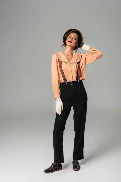 Young stylish woman touching hat while posing in retro clothes with closed eyes on grey — Stock Photo