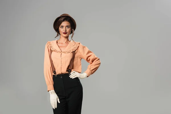 Mujer de moda en sombrero y ropa retro posando con la mano en la cadera aislado en gris - foto de stock