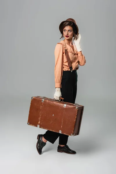 Молодая женщина в ретро-одежде ходит с винтажным чемоданом на сером — стоковое фото