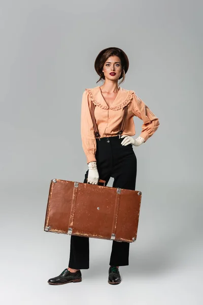 Young woman in retro clothes and hat holding vintage suitcase while posing with hand on hip on grey — Stock Photo