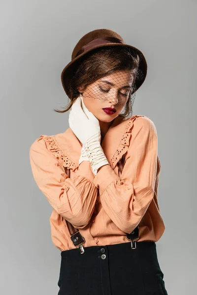 Junge Frau mit Vintage-Mütze und Handschuhen, die den Hals berühren, während sie mit geschlossenen Augen auf grau isoliert posiert — Stockfoto