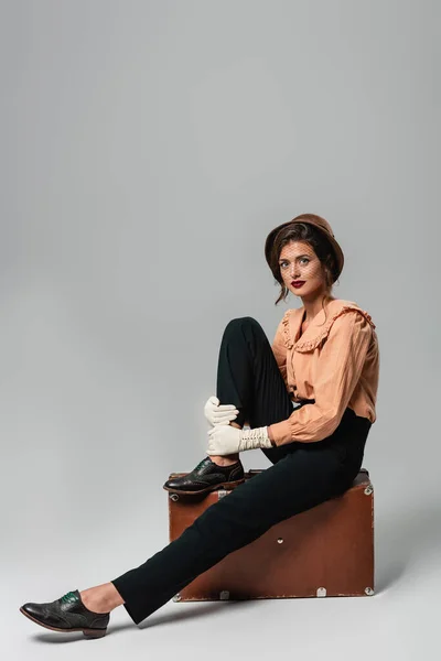 Fashionable woman in vintage clothes looking at camera while sitting on suitcase on grey — Stock Photo