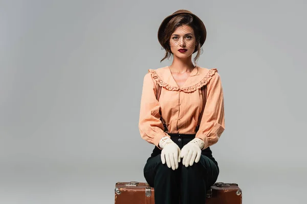 Mujer joven con sombrero y ropa retro de moda sentada en la maleta vintage en gris - foto de stock