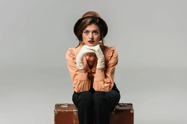 Bonita, mujer de moda sentada en la maleta vintage mientras mira a la cámara en gris - foto de stock