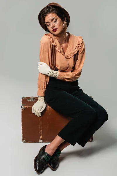 Young woman in stylish retro clothes sitting on vintage suitcase with closed eyes on grey — Stock Photo