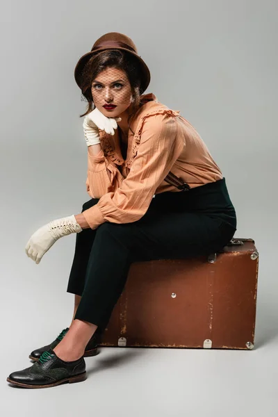 Молодая женщина в шляпе сидит на винтажном чемодане с рукой рядом лицом на сером — стоковое фото