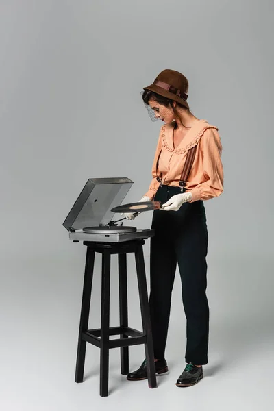 Vista completa de la mujer bonita en sombrero y ropa retro con estilo poner disco de vinilo en el tocadiscos en gris - foto de stock