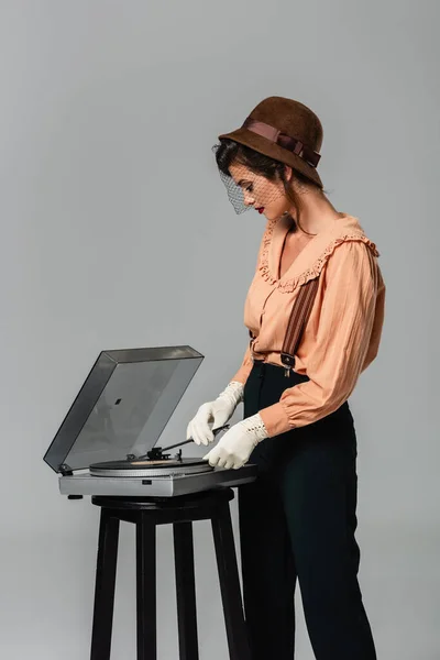 Trendy woman in retro clothes turning on record player isolated on grey — Stock Photo