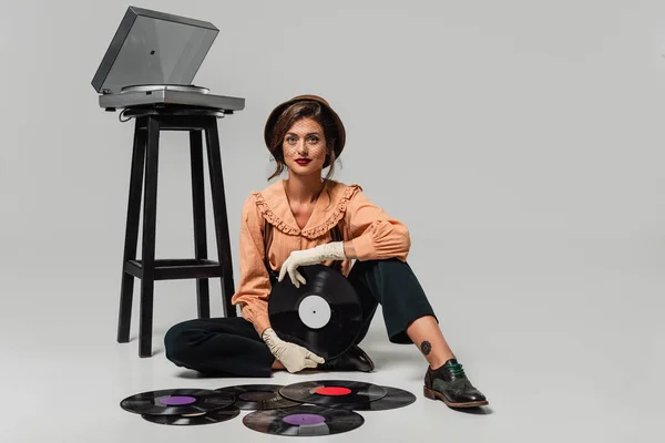 Jeune femme en vêtements rétro regardant la caméra tout en étant assis sur gris près du tourne-disque et des disques vinyle — Photo de stock