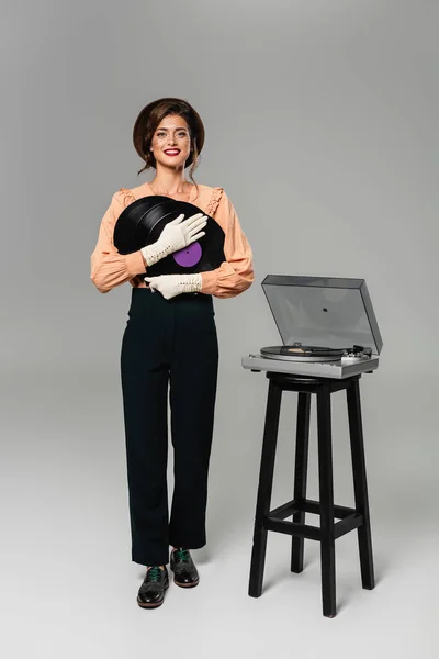 Mulher elegante feliz em roupas vintage segurando discos de vinil enquanto posando perto gravador no cinza — Fotografia de Stock