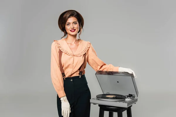 Mujer feliz en ropa retro sonriendo a la cámara mientras toca tocadiscos aislados en gris - foto de stock