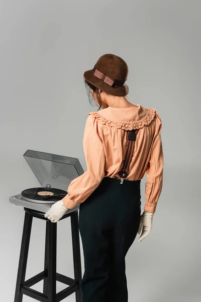 Back view of woman in hat and stylish retro clothes turning on record player on grey — Stock Photo