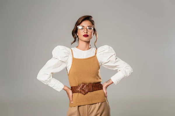 Young woman in eyeglasses and retro clothes posing with hands on hips isolated on grey — Stock Photo