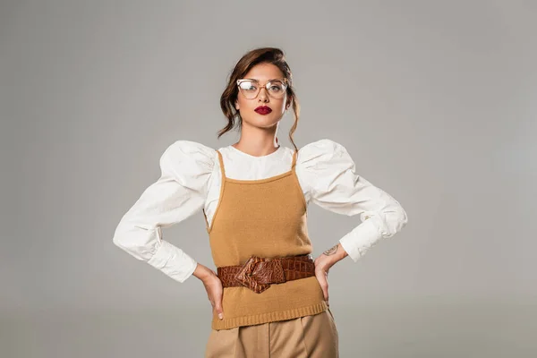 Jeune femme aux lunettes et vêtements rétro posant les mains sur les hanches isolées sur le gris — Photo de stock