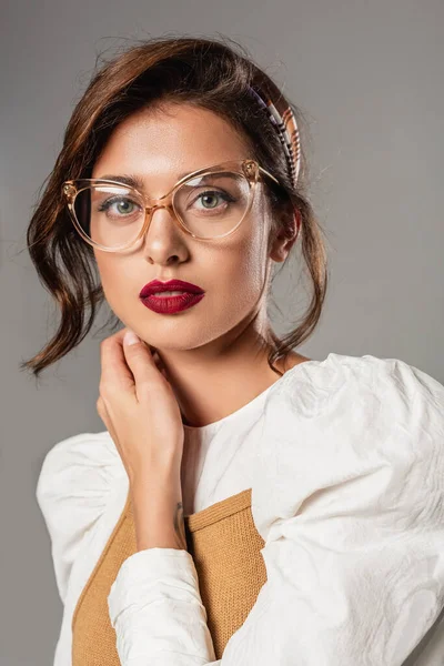 Mujer joven con gafas de vista tocando el cuello mientras mira la cámara aislada en gris - foto de stock