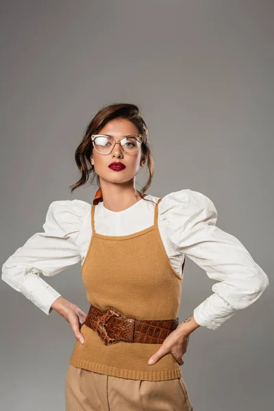 Bonita mujer en blusa blanca y chaleco beige posando con las manos en las caderas aisladas en gris - foto de stock