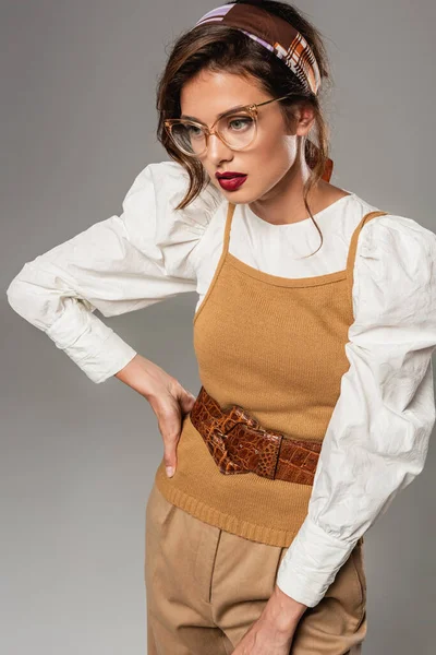 Mujer joven de moda en ropa vintage posando con la mano en la cadera aislado en gris - foto de stock