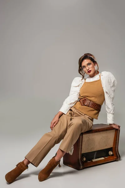 Stylish woman in vintage clothes looking at camera while sitting on retro radio receiver on grey — Stock Photo