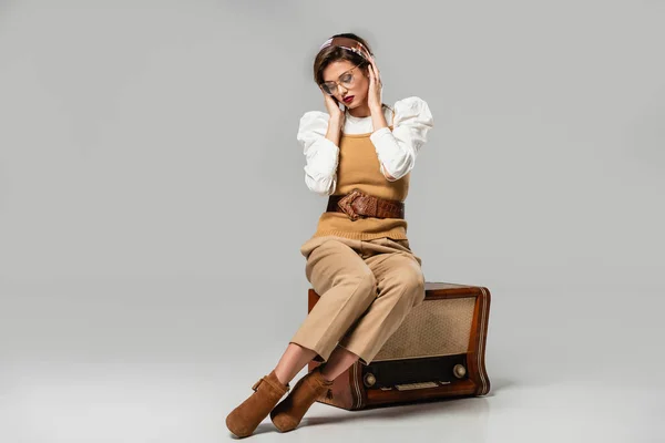 Junge Frau in trendiger Retro-Kleidung, die am Vintage-Radioempfänger auf grau sitzt und das Stirnband justiert — Stockfoto