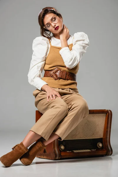 Femme à la mode touchant le cou avec les yeux fermés tout en étant assis sur un récepteur de radio rétro sur gris — Photo de stock