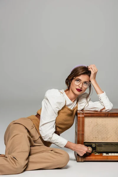 Femme à la mode s'appuyant sur un récepteur de radio vintage tout en regardant la caméra sur gris — Photo de stock