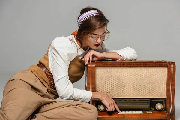 Молода жінка в модному ретро одязі натискає кнопку на старовинному радіоприймачі ізольована на сірому — стокове фото
