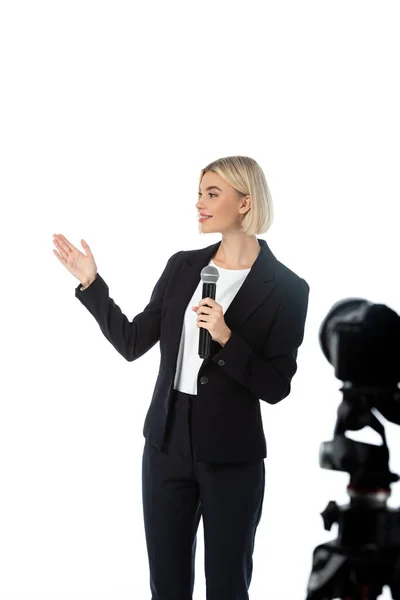 Jeune journaliste souriant avec microphone pointant avec la main près de l'appareil photo numérique sur l'avant-plan flou isolé sur blanc — Photo de stock